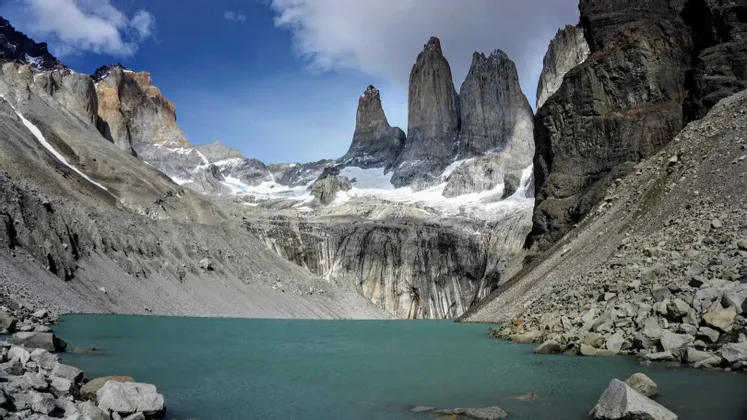 Un lago glacial turquesa se encuentra al pie de tres picos de granito escarpados y altísimos, cubiertos de nieve, bajo un cielo azul con nubes.