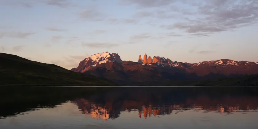 Montañas escarpadas y nevadas son iluminadas por el amanecer, con su reflejo visible en el lago en calma.