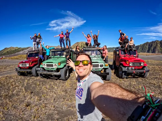 Ein Mann macht ein Selfie mit einer WeRoad-Gruppe, die in einer weiten, trockenen Landschaft unter blauem Himmel auf und um farbenfrohe Offroad-Jeeps posiert.