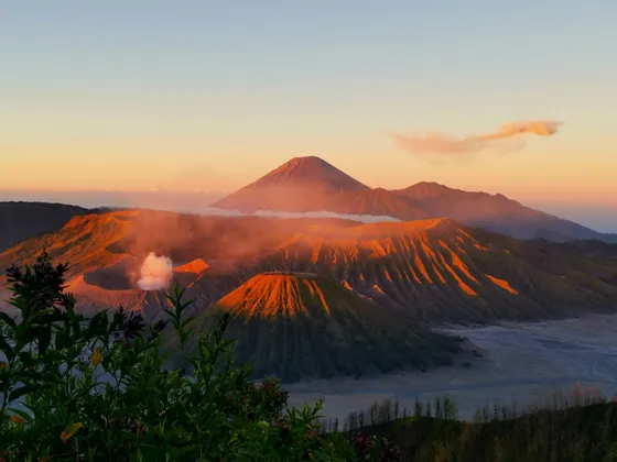 Eine Reihe vulkanischer Krater und Berge, beleuchtet vom warmen Licht des Sonnenaufgangs, wobei ein Krater eine Rauchfahne ausstößt.