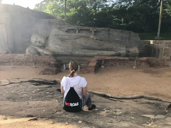 Una persona con una mochila WeRoad se sienta en una roca mirando una gran estatua de Buda reclinado tallada en un acantilado.