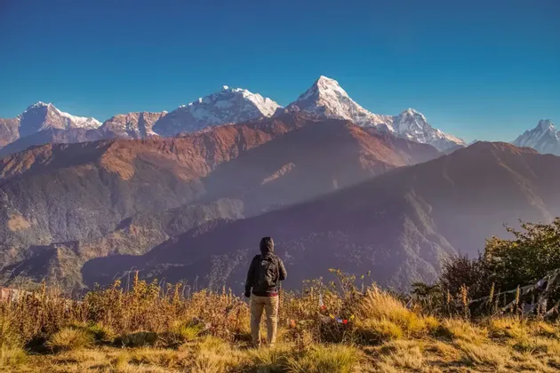 Un escursionista con uno zaino si trova su una cresta erbosa, ammirando una vista panoramica di montagne innevate sotto un cielo sereno.