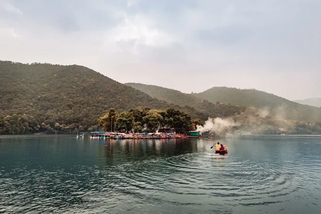Ein kleines Boot mit zwei Personen wird über einen See gerudert, auf eine bewaldete Insel zu, mit grünen Hügeln im Hintergrund.