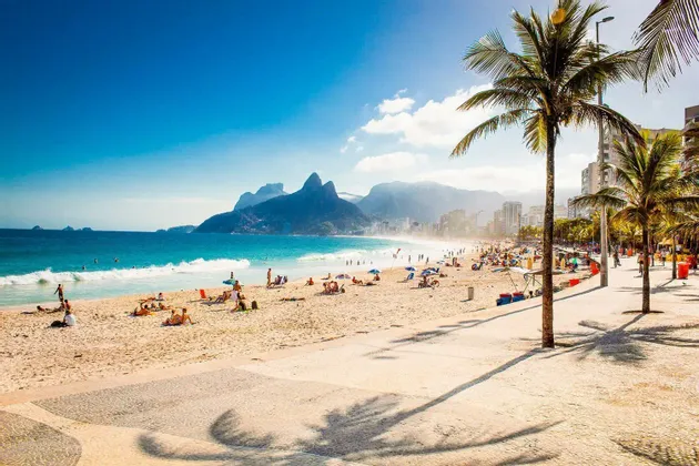 Ein belebter Sandstrand an einem sonnigen Tag, mit Palmen, einer Stadtsilhouette und Bergen im Hintergrund.
