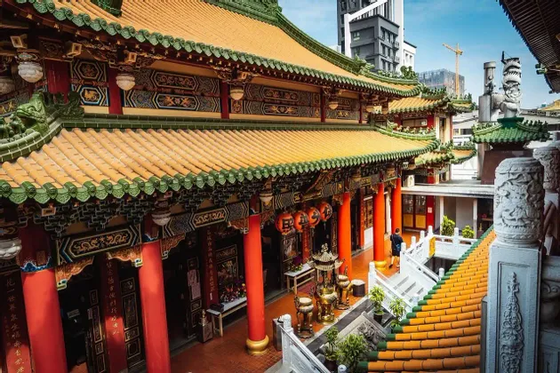Un temple traditionnel orné avec des toits de tuiles orange et des piliers rouges, sur fond de bâtiments urbains modernes.