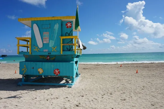 Ein türkis-gelber Rettungsschwimmerturm steht an einem Sandstrand, mit dem Ozean und einem teils bewölkten blauen Himmel im Hintergrund.