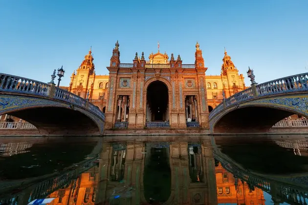 An ornate brick building with two tiled bridges is reflected in the water below during a clear sunset.