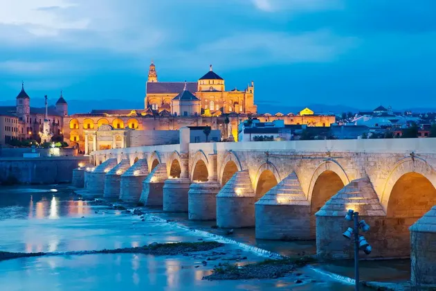 A long, multi-arched stone bridge, illuminated at dusk, crosses a river in front of a historic city skyline.