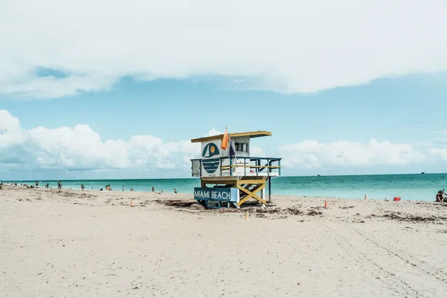 Ein bunter Rettungsschwimmerturm mit der Aufschrift 'Miami Beach' steht an einem Sandstrand mit dem Meer und einem bewölkten Himmel im Hintergrund.