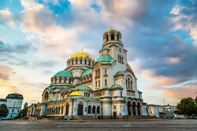 A large white cathedral with golden and green domes stands in a square under a cloudy sky at sunset.