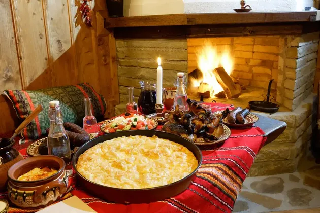 A traditional feast of savory pie, roasted meat, and salad is set on a table in front of a lit stone fireplace.