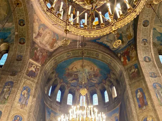 The interior of an ornate cathedral dome, covered with religious frescoes and hung with large golden chandeliers.