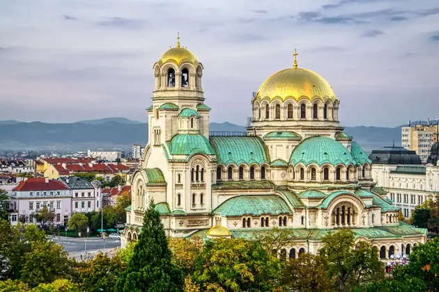 An ornate cathedral with large golden domes and teal roofs overlooks a cityscape with mountains in the background.