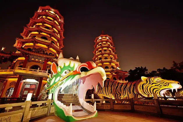 Deux grandes pagodes brillamment illuminées se découpent sur un ciel nocturne, avec d'imposantes statues colorées d'un dragon et d'un tigre au premier plan.