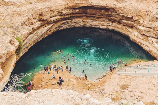 Vue plongeante sur des personnes nageant et se détendant dans l'eau turquoise d'un grand cénote entouré de falaises rocheuses.