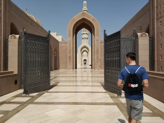 Un homme en chemise bleue et sac à dos WeRoad franchit un portail orné pour accéder à la cour ensoleillée d'une mosquée aux grandes arches.