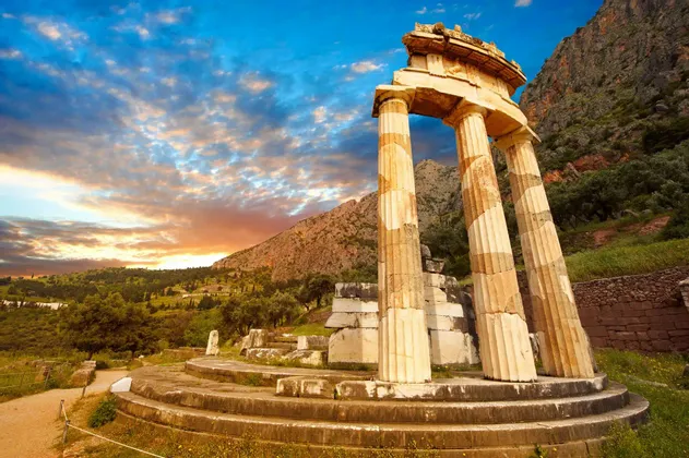 Le rovine di un antico tempio circolare con colonne sorgono su una base di pietra in una verde valle montuosa al tramonto.