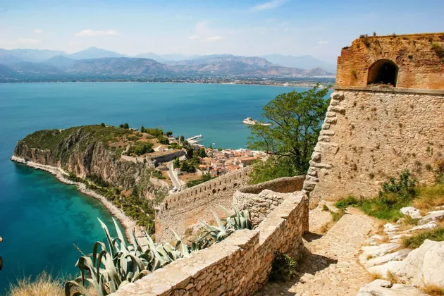 Una vista panoramica da una fortezza in pietra che domina una città costiera su una penisola, con acque turchesi e montagne lontane.