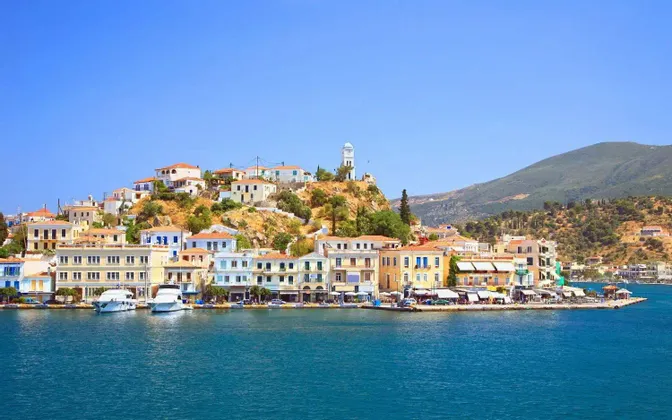 Una cittadina costiera con case colorate e una torre dell'orologio si erge su una collina sul mare azzurro brillante, con barche ormeggiate nel porto.