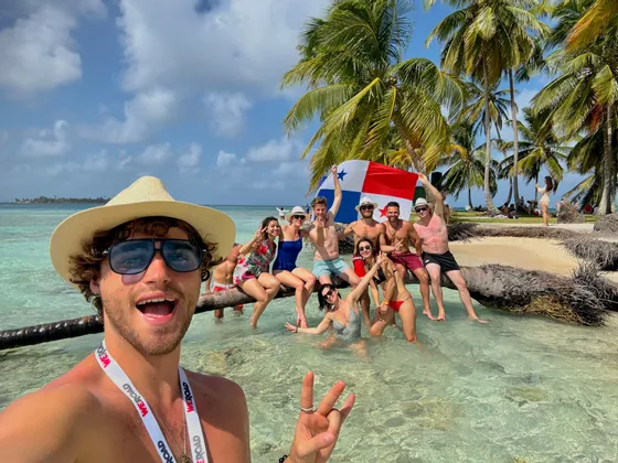 Un homme prend un selfie lors d'un voyage de groupe WeRoad sur une plage tropicale, posant sur un palmier au-dessus de l'eau et tenant un drapeau panaméen.