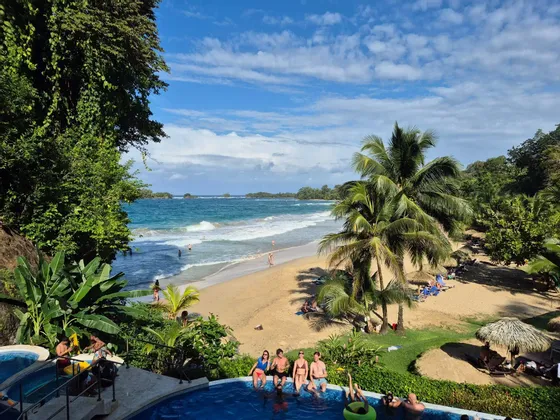 Un voyage de groupe WeRoad, détente en piscine avec vue sur une plage tropicale, ses palmiers et l'océan sous un ciel légèrement nuageux.