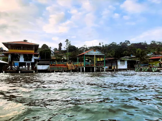 Vue depuis l'eau de maisons colorées sur pilotis sur un rivage luxuriant, l'une affichant un panneau 'Bienvenidos'.