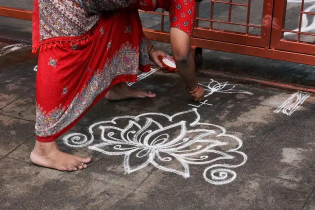 Una donna in un sari rosso a fantasia disegna un grande motivo floreale bianco sul marciapiede con della polvere.