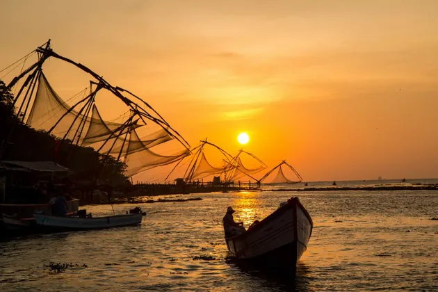Una piccola barca con una persona sull'acqua, vicino a reti da pesca cinesi stagliate contro un tramonto dorato.