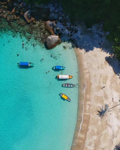 Barche in acqua turchese cristallina, spiaggia sabbiosa e bagnanti, visti dall'alto.