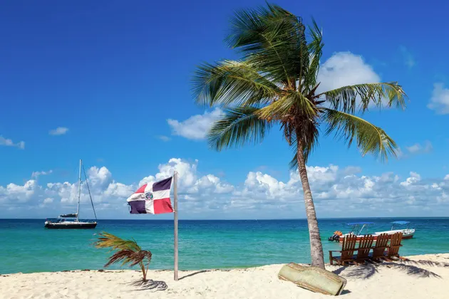 Una bandiera della Repubblica Dominicana si erge su una spiaggia di sabbia bianca accanto a una palma, con barche sul mare turchese sotto un cielo azzurro.