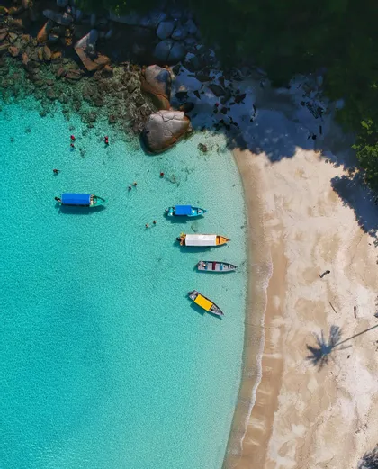Vista aerea di barche ormeggiate in acque turchesi, con persone che nuotano vicino a una spiaggia sabbiosa e scogli.