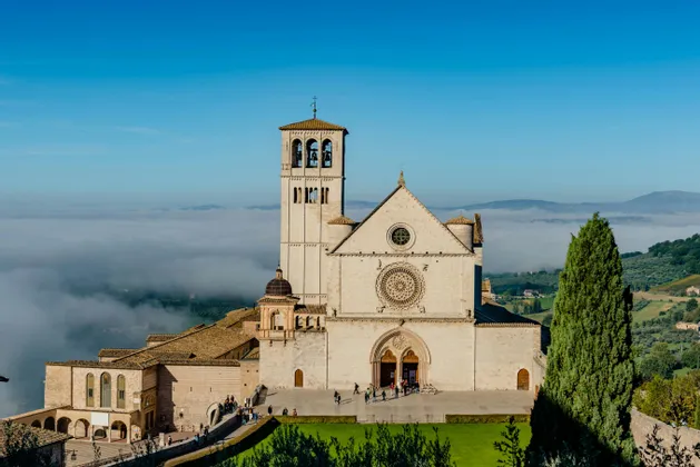 Una grande basilica di pietra con campanile si erge su un fianco di collina, dominando una valle avvolta dalle nuvole sotto un cielo azzurro e limpido.