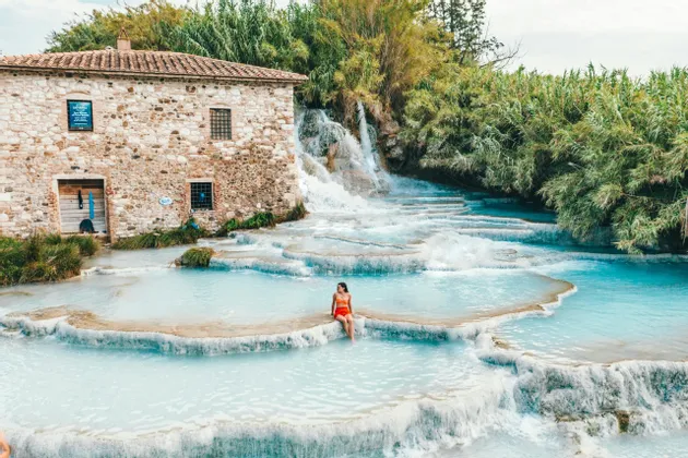 Una donna in costume arancione siede sul bordo di una piscina termale a cascata accanto a un rustico edificio in pietra.
