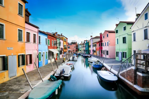 Piccole barche sono ormeggiate lungo uno stretto canale fiancheggiato da edifici dai colori vivaci sotto un cielo azzurro e limpido.