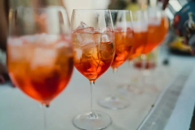 Primo piano di calici da vino con cocktail all'arancia e ghiaccio, disposti in fila su un tavolo con tovaglia bianca.