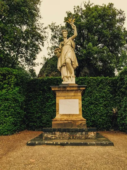Una statua classica in pietra di una donna che tiene della frutta si erge su un piedistallo in un giardino con siepi verdi.