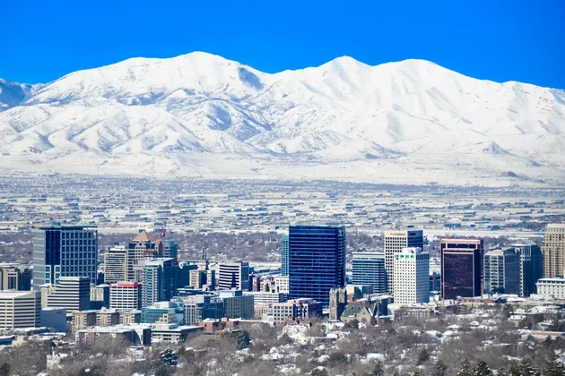 A city skyline on a clear day, with a massive snow-covered mountain range towering in the background.