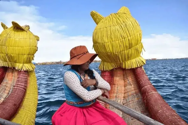 Una donna con un cappello marrone e abiti tradizionali colorati siede su una barca di giunchi con teste decorative a forma di gatto, galleggiando sull'acqua blu.
