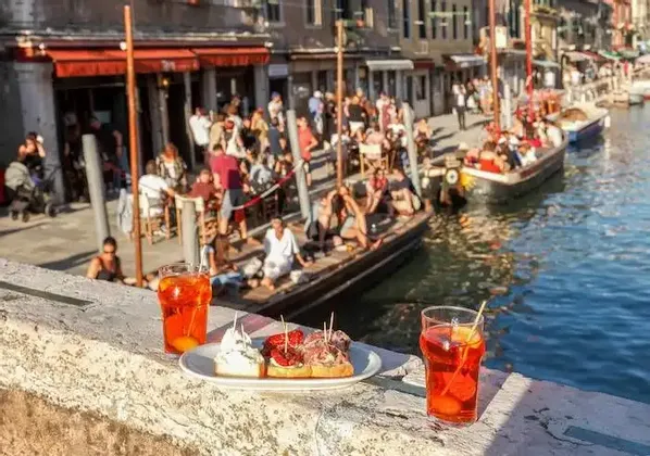 Due cocktail arancioni e stuzzichini su un davanzale in pietra, con vista su un vivace canale dove gente socializza su barche e in un caffè sul lungomare.