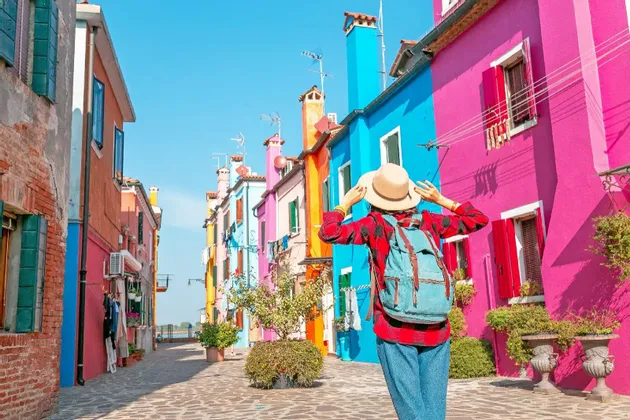 Una persona con uno zaino, vista da dietro, si tiene il cappello mentre è in piedi su una strada fiancheggiata da edifici colorati.