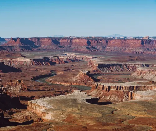 An expansive view of a deep red rock canyon with a river snaking through the canyon floor under a clear sky.