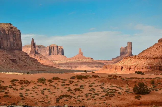 A wide shot of a red sand desert valley with large sandstone buttes and mesas rising in the distance under a blue sky.