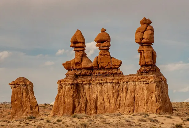 Three large, reddish-brown hoodoo rock formations stand in a desert landscape under a partly cloudy sky.