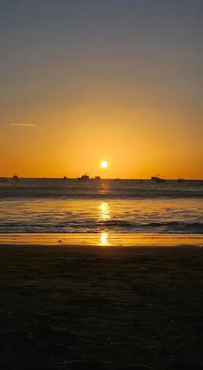 Die Sonne versinkt über dem Meer, Boote zeichnen sich als Silhouetten am Horizont ab, während sich ihr Schein auf dem nassen Sand spiegelt.