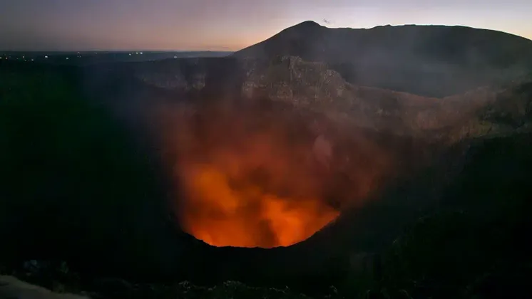 Weitblick auf einen Vulkankrater bei Sonnenuntergang, mit glühender orangefarbener Lava und aufsteigendem Rauch.