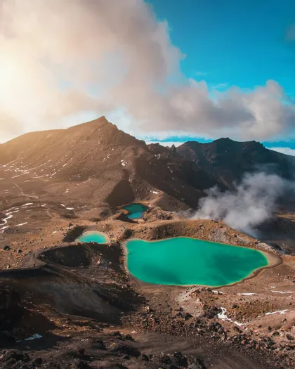 An aerial view of three turquoise crater lakes nestled in a rocky, volcanic landscape with steam rising from the ground.