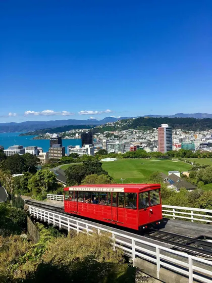 A red cable car travels on a track overlooking a coastal city with a large green park and a bay under a clear blue sky.