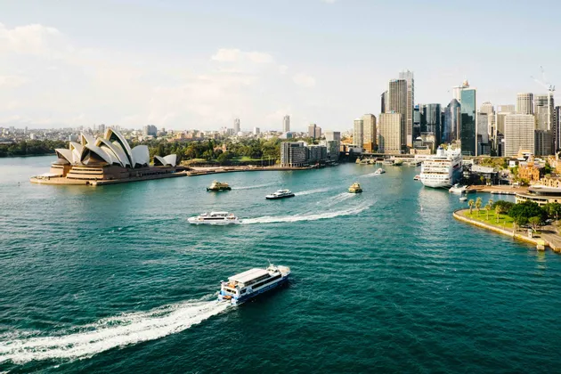 An aerial view of a busy harbor with ferries, a large opera house, and a modern city skyline under a clear sky.