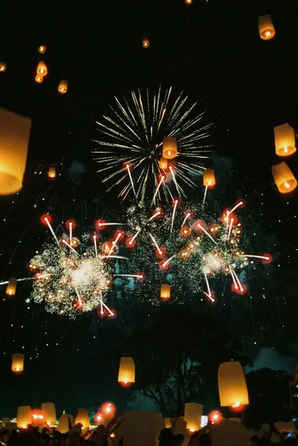 Fuochi d'artificio esplodono in un cielo notturno buio, con numerose lanterne di carta illuminate che si librano verso l'alto da una folla sottostante.