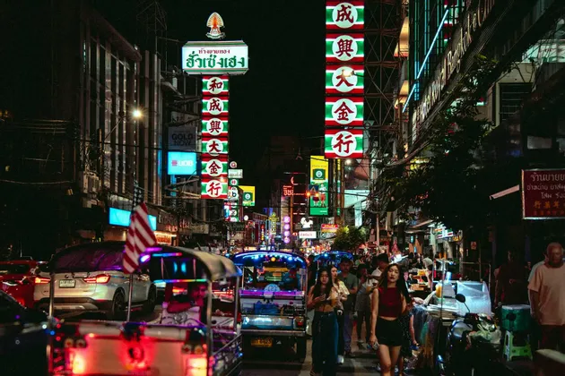 Una strada cittadina affollata di notte con persone che camminano e tuk-tuk nel traffico, illuminata da insegne al neon colorate con caratteri asiatici.